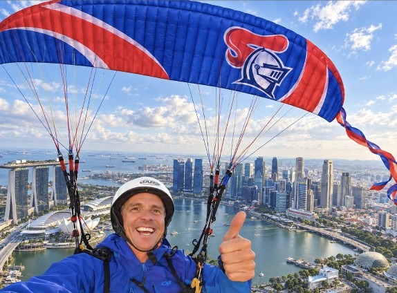 A paraglider under a red and blue Knights canopy giving a thumbs up above the Singapore skyline.