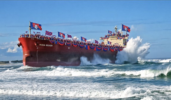 A red bulk carrier flying Knights flags pushing through heavy surf.
